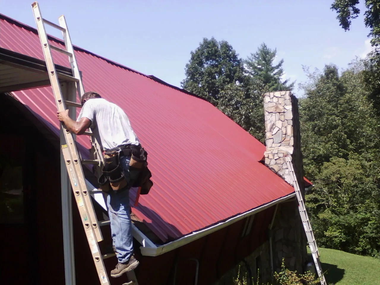 Roofer on a ladder beside a red metal roof and stone chimney — Soffit & Fascia Repair by Roofing Brothers in Petrolia